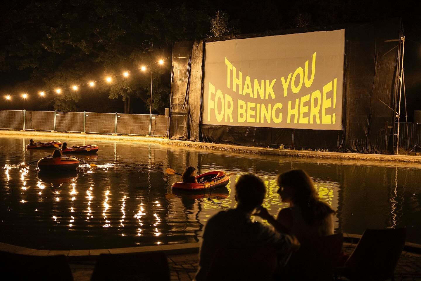 An open-air cinema at night: people in inflatable boats on a pool, and a couple on the shore, watching a screen that reads “Thank you for being here!”